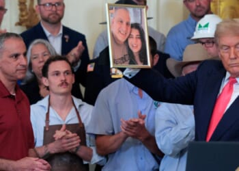 Joe Abraham, left, in red shirt, watches as President Donald Trump holds up a photograph of Abraham and his daughter, Katie Braham, during an event in the East Room of the White House in June 2025.