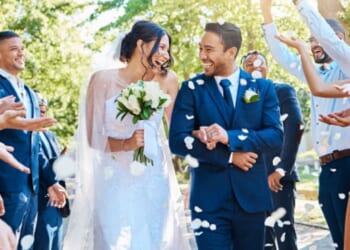 A couple gets married outdoors as attendees throw flower petals on them.