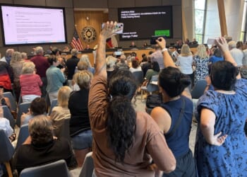 A large crowd attends a public meeting in a modern room; many people are standing, some with raised fists, facing officials seated at a long front desk with digital displays and American flags.