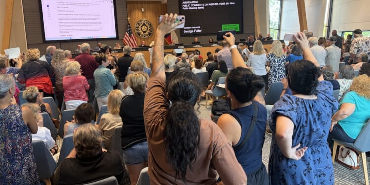 A large crowd attends a public meeting in a modern room; many people are standing, some with raised fists, facing officials seated at a long front desk with digital displays and American flags.