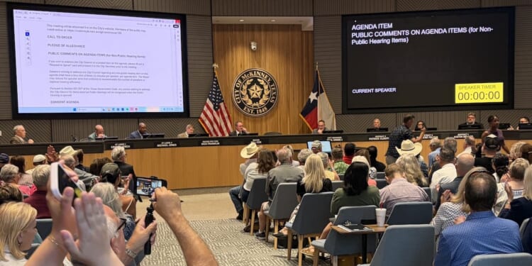 A large group attends a city council meeting. People sit facing a panel of officials at the front. Digital screens display agenda items and a timer. U.S. and Texas flags are visible behind the officials.