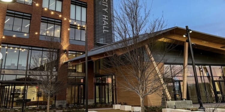 Modern city hall building with large windows, string lights, and landscaping with bare trees at dusk; CITY HALL sign is visible on the buildings exterior.