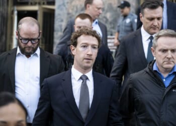 Mark Zuckerberg in a suit walks outside with several other men, some in suits and one in a jacket, all appearing serious. They are leaving a building with a stone facade; a uniformed guard is in the background.