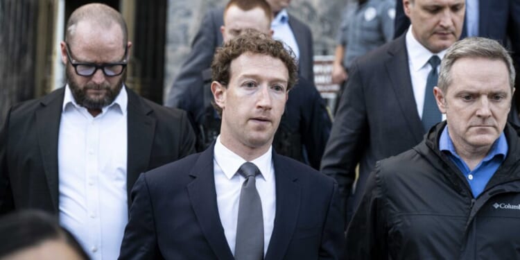 Mark Zuckerberg in a suit walks outside with several other men, some in suits and one in a jacket, all appearing serious. They are leaving a building with a stone facade; a uniformed guard is in the background.