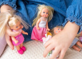 A young girl plays with female toy dolls on her bed.