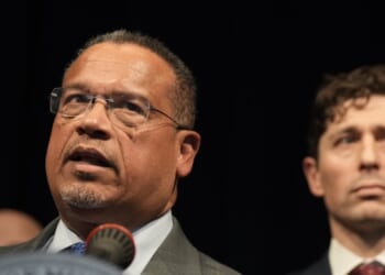 A man wearing glasses and a suit speaks at a microphone, with another man in a suit standing slightly behind him against a dark background.