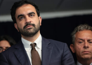 Zohran Mamdani in a blue suit and brown tie stands in front of a dark background, looking serious, Two other people, a woman and a man, are partially visible behind him.