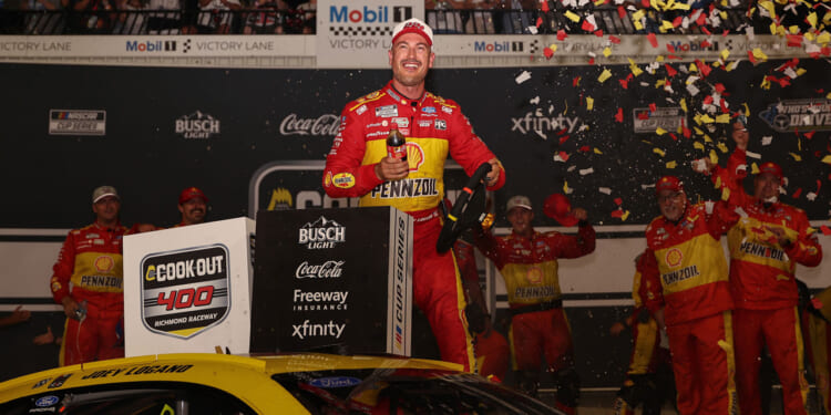 NASCAR race car driver Joey Logano in a yellow Pennzoil suit celebrates atop his car in victory lane as confetti falls. Crew members behind him cheer and clap, while racing sponsor logos cover the background.