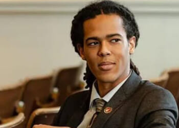 Democrat Jonah Wheeler is not afraid to buck his party on transgender orthodoxy and gender ideology. Pictured: Wheeler, a man with long, dark hair in dreadlocks, wearing a suit and tie, sits in an auditorium with brown seats, looking to the side.