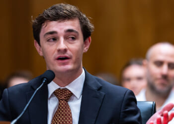 Nick Shirley, a young man in a suit and tie, speaks into a microphone at a public hearing, with people seated and listening in the blurred background.