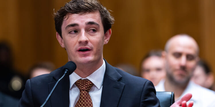 Nick Shirley, a young man in a suit and tie, speaks into a microphone at a public hearing, with people seated and listening in the blurred background.