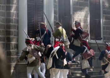 Men in historical military uniforms, carrying muskets and bags, ascend stone steps toward a building with closed shutters and columns, suggesting a reenactment of a historical event.