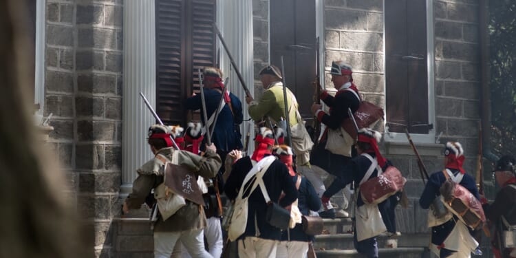 Men in historical military uniforms, carrying muskets and bags, ascend stone steps toward a building with closed shutters and columns, suggesting a reenactment of a historical event.