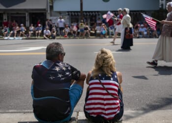A man and woman, both wearing American flag-themed clothing, sit on the grass watching a parade with people carrying U.S. flags on the street. Spectators line the sidewalk in the background.