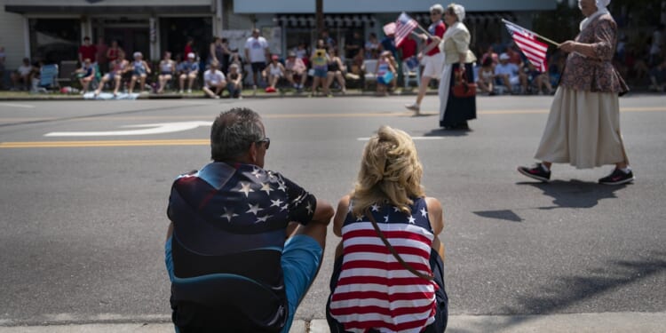 A man and woman, both wearing American flag-themed clothing, sit on the grass watching a parade with people carrying U.S. flags on the street. Spectators line the sidewalk in the background.