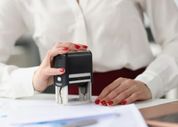 A person in a white shirt stamps a document on a desk, with red-painted nails visible. Papers and a laptop are also on the desk.