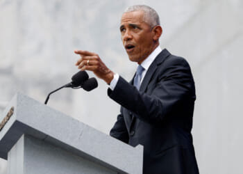 Barack Obama in a dark suit and tie, speaks at a gray podium with microphones, gesturing with his right hand against a light stone background.