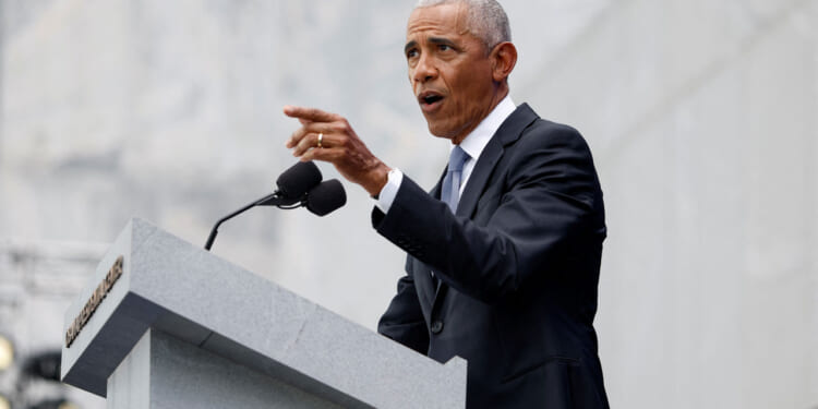 Barack Obama in a dark suit and tie, speaks at a gray podium with microphones, gesturing with his right hand against a light stone background.