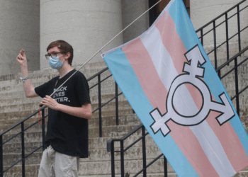 A person wearing a mask and a Black Lives Matter shirt stands on stone steps in Ohio, holding a large transgender pride flag next to black railings.