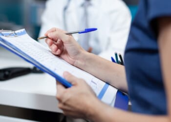 A person holding a clipboard writes notes with a pen, while another person in a white coat is blurred in the background, suggesting a medical or healthcare setting.