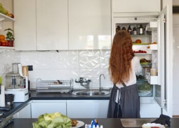 A woman organizing her refrigerator filled with fresh fruits and vegetables in a modern kitchen.