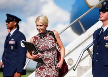 Trump Aide Natalie Harp holds a folder and exits an airplane, flanked by two uniformed Air Force members standing guard. The sky is blue with clouds in the background.