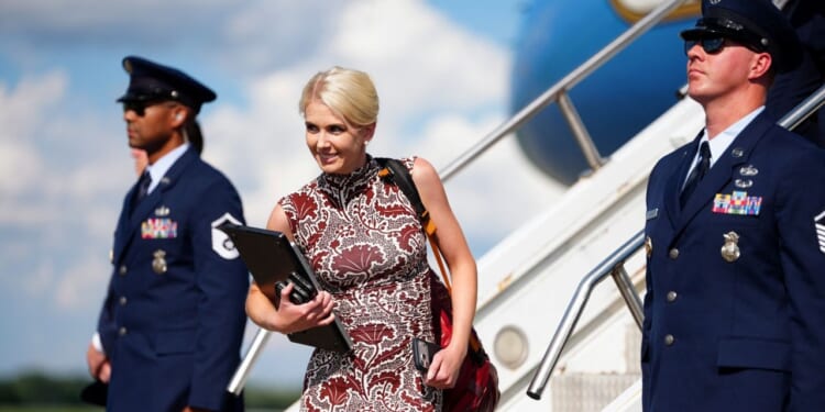Trump Aide Natalie Harp holds a folder and exits an airplane, flanked by two uniformed Air Force members standing guard. The sky is blue with clouds in the background.