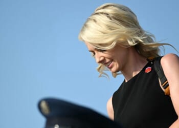 Natalie Harp, a top Trump aide, appears with blonde hair, wearing a sleeveless black top and backpack. She looks down and smiles against a clear blue sky.