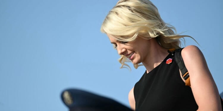 Natalie Harp, a top Trump aide, appears with blonde hair, wearing a sleeveless black top and backpack. She looks down and smiles against a clear blue sky.