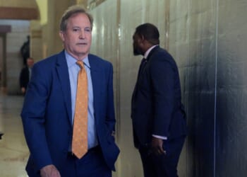 Ken Paxton in a blue suit with an orange polka dot tie walks in a hallway, while another man in a dark suit stands nearby, facing away.