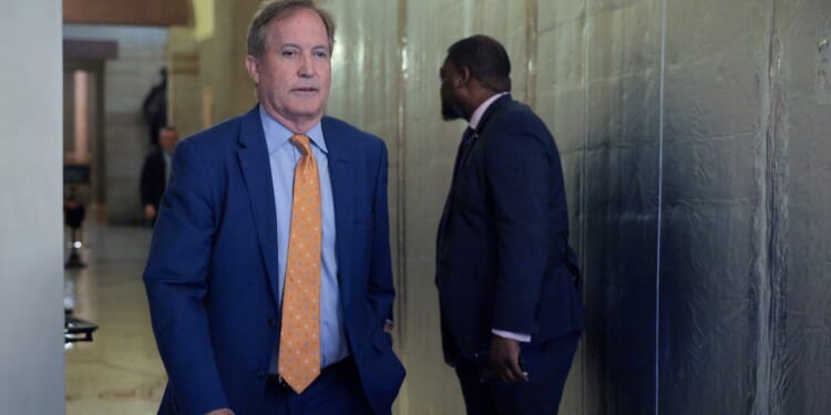 Ken Paxton in a blue suit with an orange polka dot tie walks in a hallway, while another man in a dark suit stands nearby, facing away.