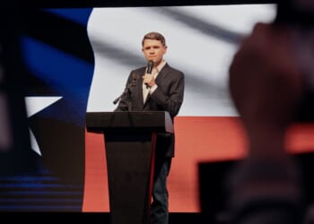 A man in a suit speaks at a podium on stage, with a large Texas flag projected behind him and blurred audience signs in the foreground.