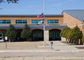 A brick school building with two flags at half-staff in front, surrounded by trimmed trees and a sidewalk under a clear blue sky.