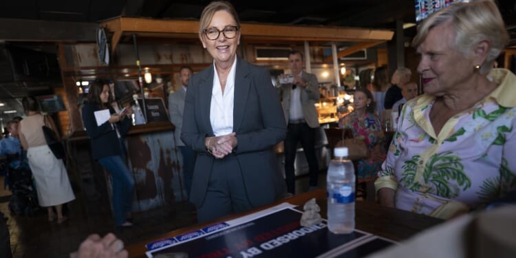 A woman in a suit smiles while standing near a table with a campaign sign and water bottle. People sit and mingle in a casual indoor setting with warm lighting.