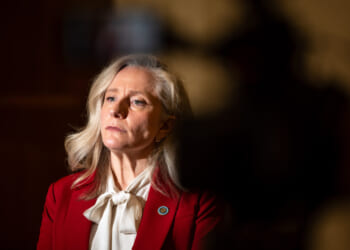 Abigail Spanberger with long gray hair, who wears a red blazer and white blouse, looks slightly to the side. The background is dimly lit with a dark, shadowy area on the right.