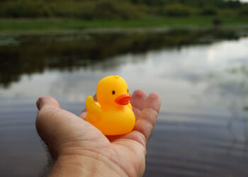 A person holds a small yellow rubber duck over calm water, with greenery and cloudy sky reflected on the lakes surface in the background.