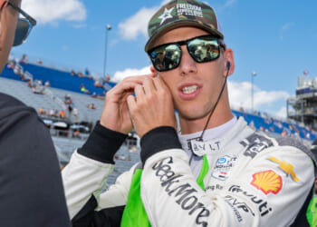 A race car driver in a white and green suit adjusts his earpiece while talking to another person at a racetrack. Blue sky and grandstands with spectators are visible in the background.