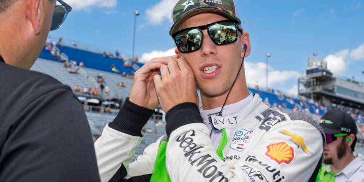 A race car driver in a white and green suit adjusts his earpiece while talking to another person at a racetrack. Blue sky and grandstands with spectators are visible in the background.