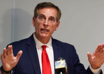 A man in a navy suit, red tie, and glasses speaks at a podium, gesturing with both hands raised. A microphone with a yellow and blue 2 logo is visible in front of him against a plain background.
