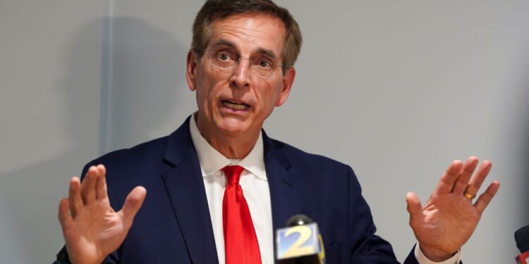 A man in a navy suit, red tie, and glasses speaks at a podium, gesturing with both hands raised. A microphone with a yellow and blue 2 logo is visible in front of him against a plain background.