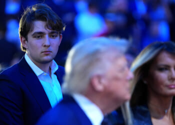 Barron Trump in a suit looks serious while standing behind his parents in focus. The background is filled with people and blue lighting.
