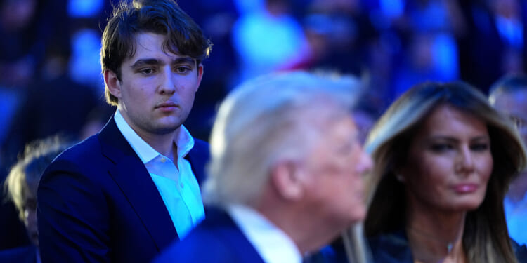 Barron Trump in a suit looks serious while standing behind his parents in focus. The background is filled with people and blue lighting.