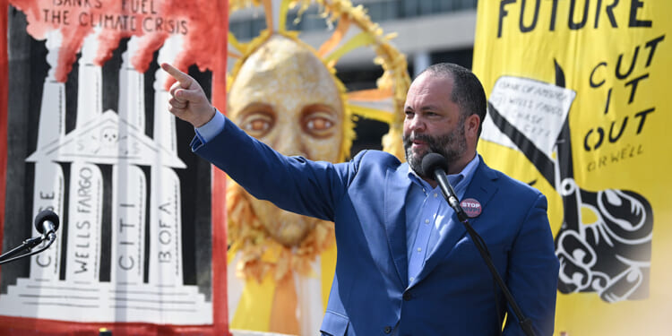 A new report highlights foreign funding for climate activist groups. Pictured: Sierra Club Executive Director Ben Jealous, a man in a blue suit speaks into a microphone and points, with climate protest signs and a sun mask in the background. Signs criticize banks and call for action against climate change.
