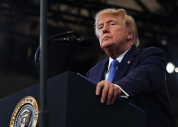 President Donald J. Trump wears a dark suit and blue tie stands at a podium with a presidential seal, speaking into a microphone in a well-lit room.