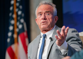 A man in a gray suit gestures while speaking onstage, with an American flag and a blue backdrop visible behind him.