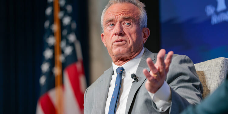 A man in a gray suit gestures while speaking onstage, with an American flag and a blue backdrop visible behind him.