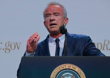 A man in a blue suit speaks at a podium with the U.S. presidential seal, gesturing with his right hand. The background shows partial text ending in Rx.gov..