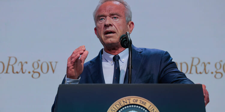 A man in a blue suit speaks at a podium with the U.S. presidential seal, gesturing with his right hand. The background shows partial text ending in Rx.gov..