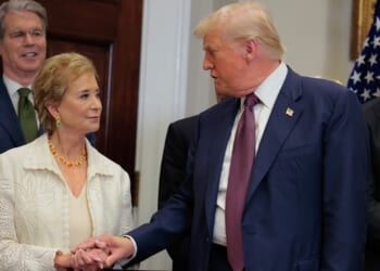 A woman and a man in formal attire shake hands in a room with other people and an American flag in the background.