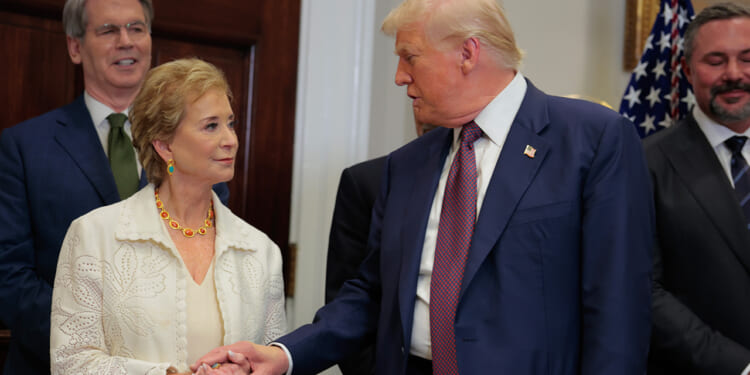 A woman and a man in formal attire shake hands in a room with other people and an American flag in the background.